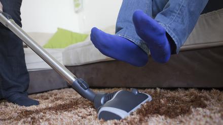 Ein Mann hebt seine Beine, während eine Frau den Teppich saugt.
