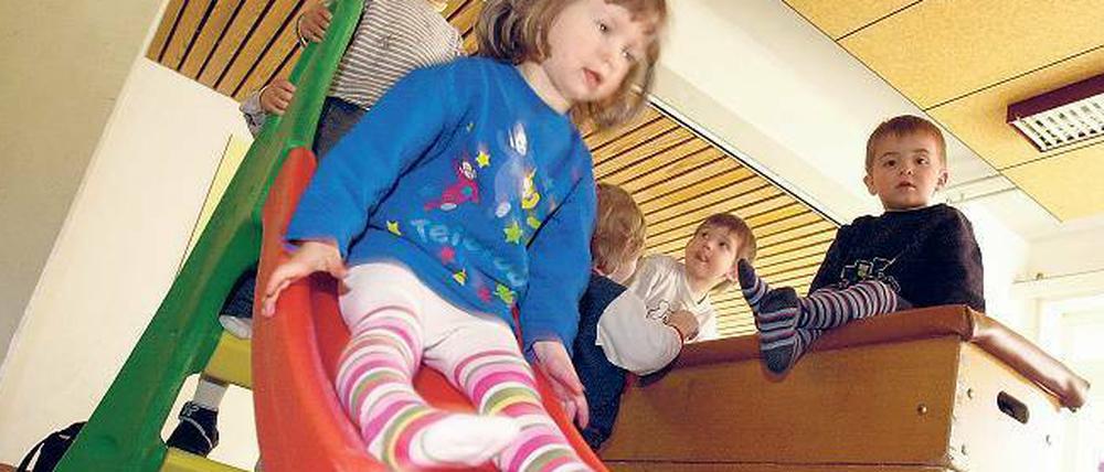 Ein kleines Mädchen rutscht im Innenraum einer Kitag von einer Plastikrutsche, andere Kinder turnen auf einem Kasten.