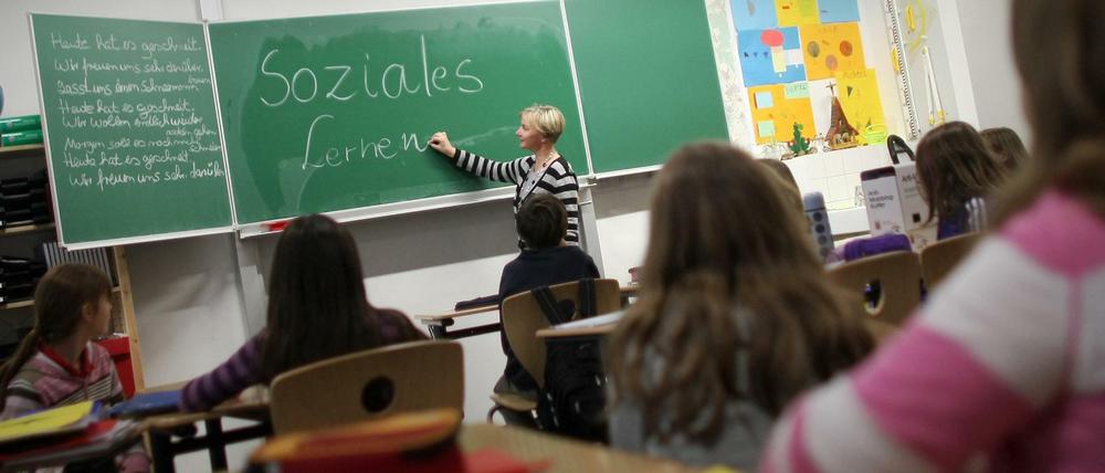 Eine Lehrerin schreibt "Soziales Lernen" an eine Tafel, im Klassenraum sitzen etwa zehnjährige Kinder.